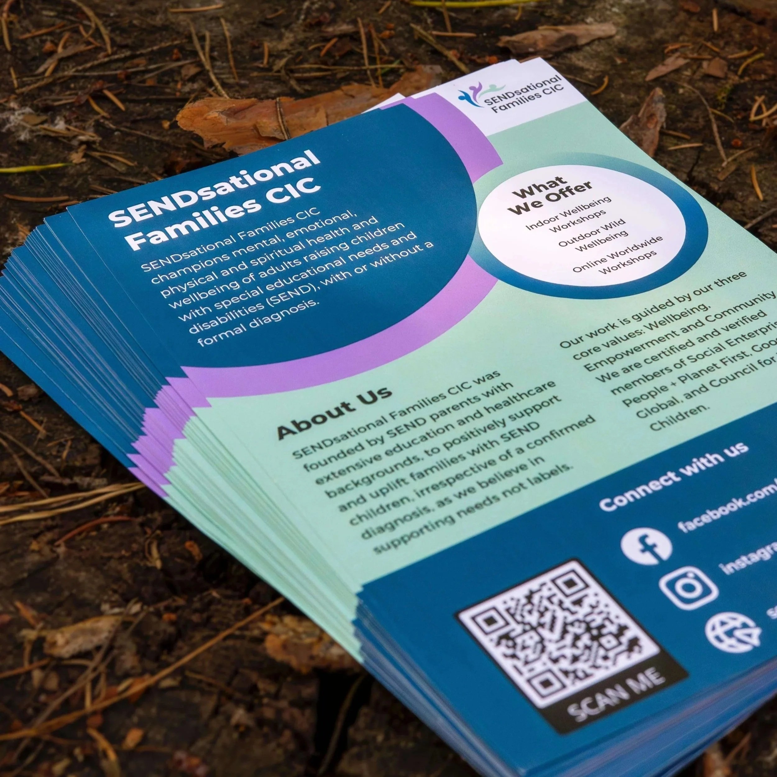 A stack of colorful flyers for SENDsational Families CIC, placed on the ground among dirt and fallen leaves. The flyer includes information about the organization, their services, and contact details, with QR code and social media icons.