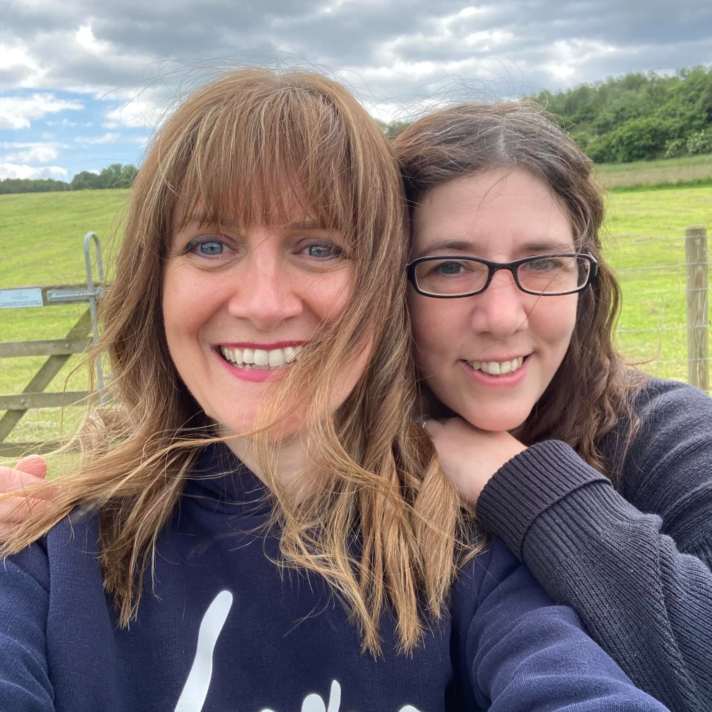 Two women taking a selfie outdoors in a green field with a cloudy sky.